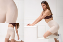 A model wearing beige compression shorts and a matching ribbed waistband top poses on white display blocks in a bright studio setting.